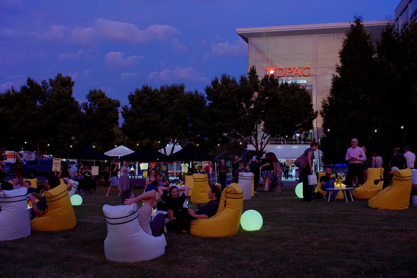 Guests relaxing in yellow and white high-back bean bag chairs with glowing globes on the DPAC lawn at dusk, the DPAC sign lit behind them, at Discover Durham's Good Moon event